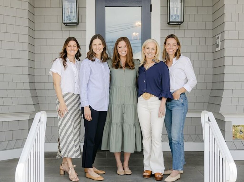 Five women stand smiling in front of a gray shingled house with a black door, posing on a porch.