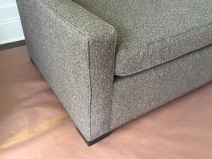 A close-up image of the corner of a gray upholstered couch with one armrest visible, placed on a cardboard floor covering.