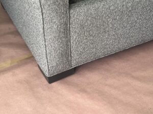 Close-up of the corner of a gray upholstered sofa with a black square leg, placed on a taupe-colored floor.