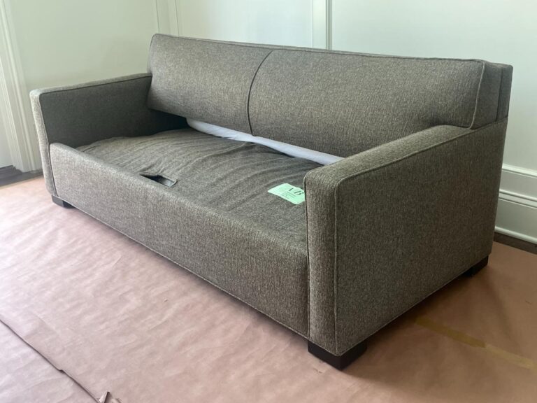 A brown fabric sofa with a slightly indented seat cushion and a green tag on the right armrest, positioned in a room with white walls and covered flooring.