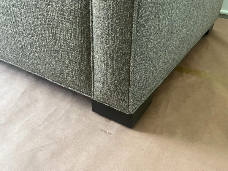 A close-up of the bottom corner of a gray fabric-covered sofa with black wooden legs on a paper-covered floor.