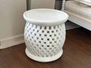 A white woven ceramic side table sits on a dark wood floor near a chair with cushions.