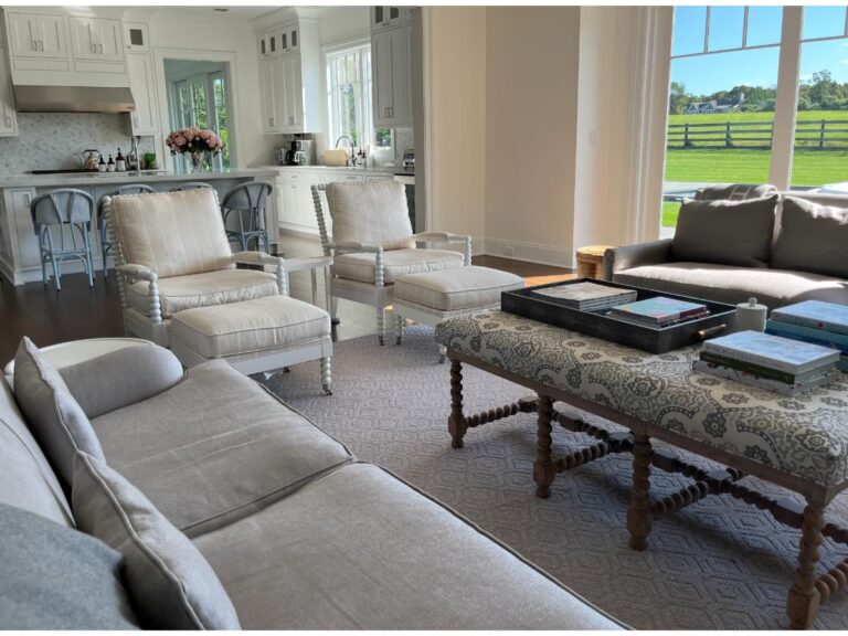 A bright living room with beige sofas, armchairs, a patterned ottoman, and a coffee table with books. The room connects to an open kitchen and features large windows overlooking a green field.