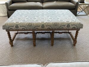 A wooden coffee table with turned legs features a patterned cushion on top. It is placed on a patterned carpet with a gray sofa set and a small side table in the background.