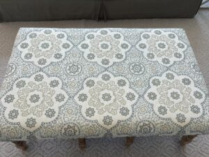 A patterned ottoman with floral and geometric designs in neutral tones on a light background. The ottoman sits on a carpeted floor.