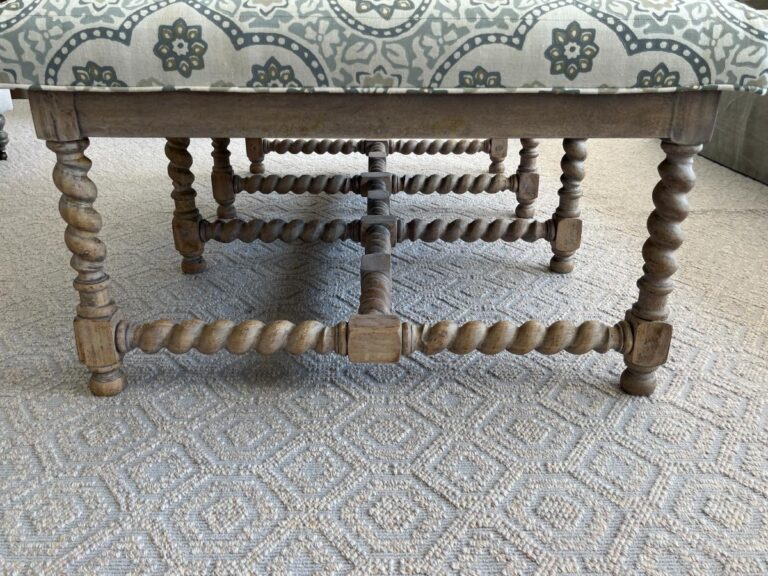 Close-up view of a wooden bench with intricately carved legs and a patterned upholstered seat on a carpeted floor.