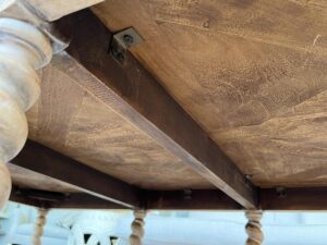 Underneath view of a wooden table showing the tabletop slats, wooden beams for support, and intricately carved table legs. The underside is detailed with metal hardware.