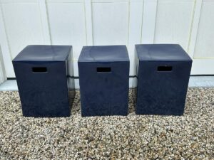 Three black, cube-shaped stools with handle cutouts are arranged in a row on a gravel surface against a white wall.