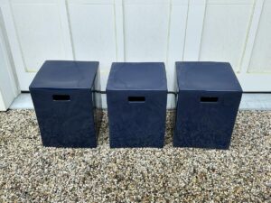 Three dark blue cubic stools with handle cutouts are placed on a gravel surface in front of a white paneled door.