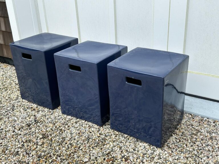 Three square, dark blue, glossy stools with handle cutouts sit in a row on pebbled ground against a white wall.