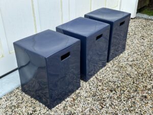 Three dark blue rectangular stools with handle cutouts on a gravel surface in front of a white wall.