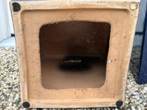 Square bottom view of an empty, tan ceramic planter with small black pads on each corner and a central drainage hole. The surface is rough with visible wear and minor discoloration.