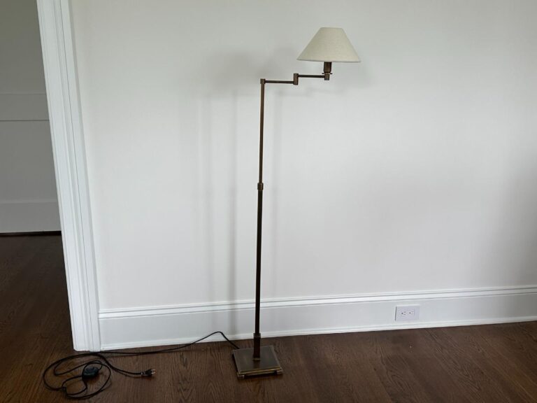 A brass floor lamp with an adjustable arm and a small white lampshade stands against a plain white wall. The cord is visible and plugged into an outlet at the base of the wall.