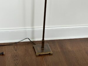 A floor lamp with a square brass base stands on a wooden floor near a white wall, with an exposed black power cord.