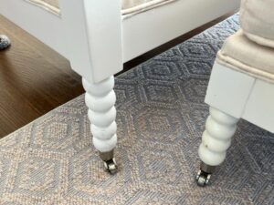 Close-up of white chair legs with casters on a patterned rug. The chair legs feature a spindle design. The rug has a geometric pattern in beige and gray tones.