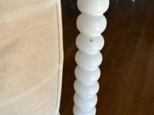 Close-up of a white, round, wooden leg of a piece of furniture with a small hole, next to a beige, upholstered surface.