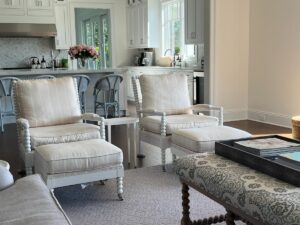 A modern living area features two beige armchairs with matching ottomans, a patterned coffee table, and a view into a white kitchen with bar stools and flowers on the counter.