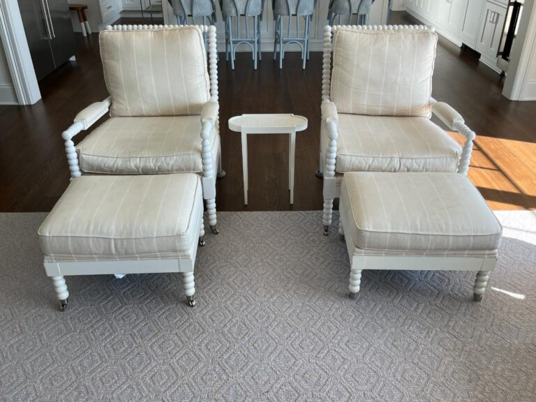 Two beige armchairs with matching ottomans are placed in front of a small side table in a well-lit, modern living space with wooden floors.