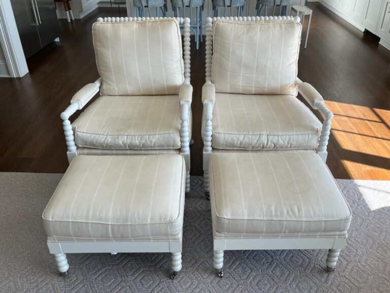 Two identical beige armchairs with matching ottomans in a living room setting. The furniture features wooden spindle legs and a striped upholstery pattern.