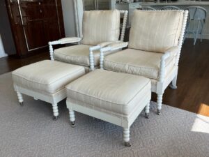 Two cream-colored, cushioned armchairs with matching ottomans sit on a light-colored carpet in a living room with wooden flooring and cabinetry in the background.