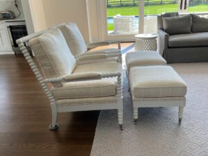 Two upholstered armchairs with matching ottomans, each featuring a white frame and spool detailing, are arranged in a living room setting with a view of the outdoors.