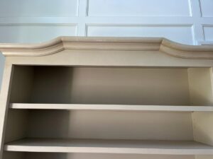 A light-colored, empty bookshelf with a decorative top molding, viewed from below. The background shows white paneled walls.