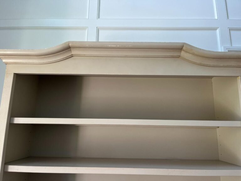 A light-colored, empty bookshelf with a decorative top molding, viewed from below. The background shows white paneled walls.