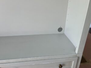 A white built-in shelf with a smooth surface, featuring a small circular hole on the back panel. There's a drawer below the shelf, partially showing a brass knob.