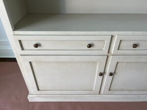 A white wooden cabinet with two drawers and two doors, featuring round brass knobs, standing in an unoccupied space.