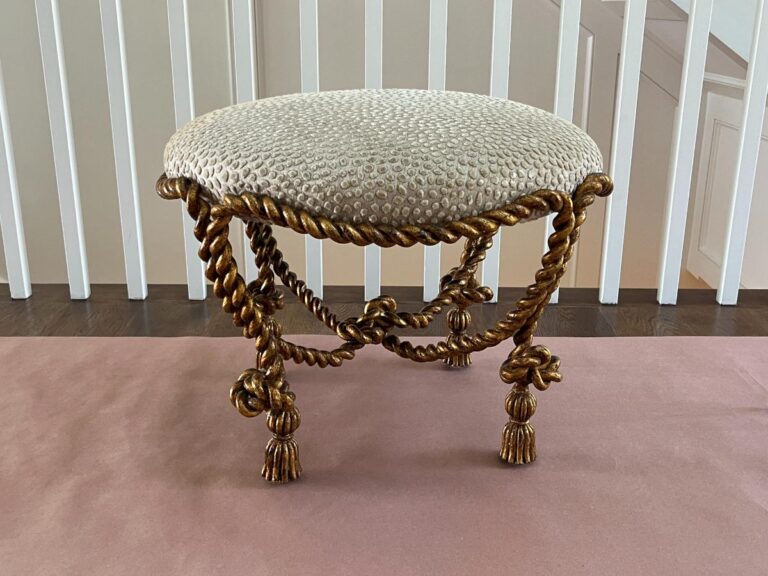 A round upholstered stool with a beige, textured top, and elaborate, intertwined golden rope legs, placed on a light pink surface in front of white vertical slats.