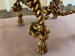 Close-up of ornate, twisted gold-colored table legs designed to look like thick rope knots, with tasseled ends. The surface beneath the legs appears to be a brown paper sheet.