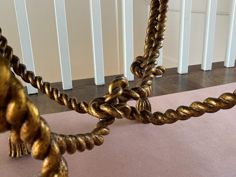 A close-up view of a gold-colored, twisted rope-style metal table leg on a light pink surface, with white vertical rails in the background.