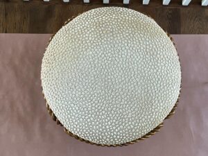 Top view of a beige, textured circular cushion with a braided trim, placed on a pink mat, against a wooden floor background.