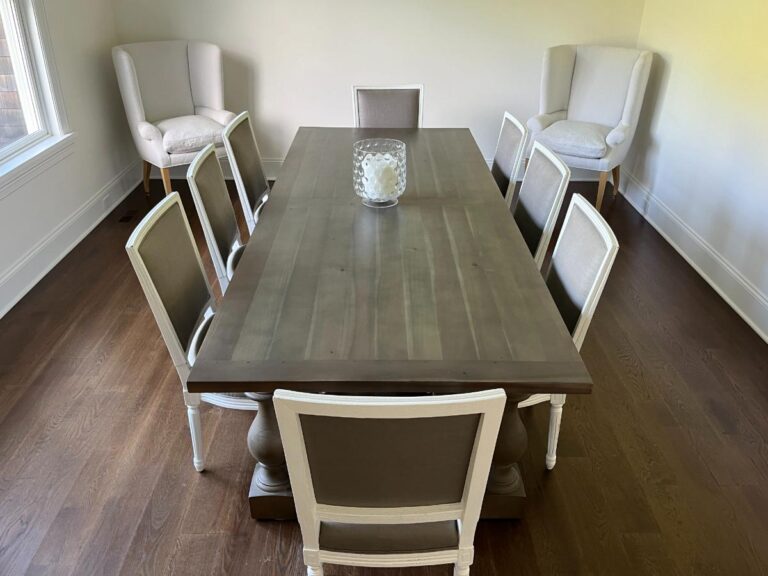 A wooden dining table with eight chairs, six chairs around the table and two armchairs in the corners of the room. The table has a glass vase in the center and the room has wooden flooring.