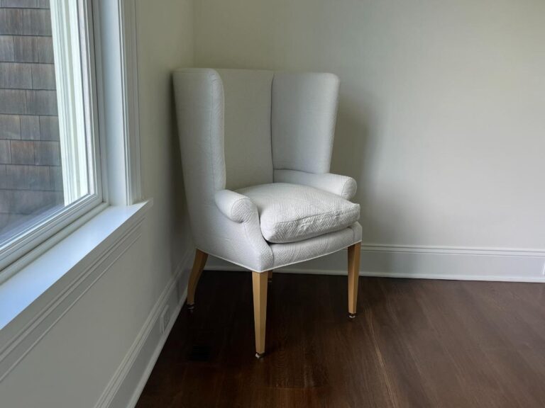 A white wingback chair with wooden legs is positioned in a corner of a room with white walls and a wooden floor. A window is visible to the left of the chair, letting in natural light.