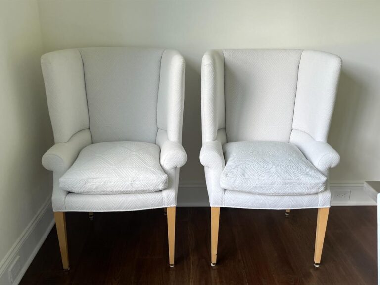 Two white upholstered armchairs with light wood legs are placed side by side against a plain wall on a dark wooden floor.