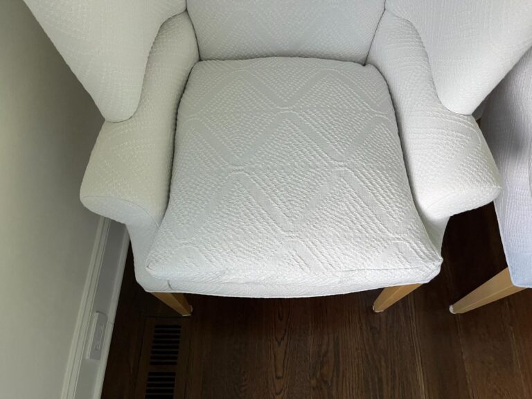 White upholstered armchair with a diamond-patterned fabric, slightly angled to the left of the image, placed on a dark wooden floor near a wall vent.