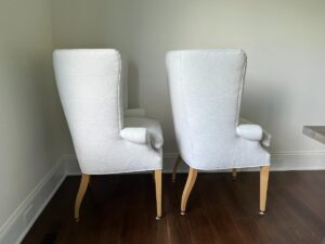 Two white, upholstered armchairs with light wooden legs are positioned side by side on a hardwood floor against a plain white wall.