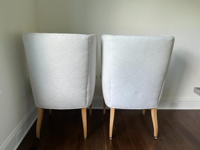 Two white armchairs with a diamond-patterned fabric and wooden legs are placed side by side, facing away from the camera against a light-colored wall.