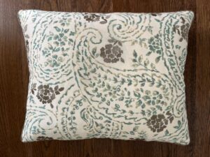 A square cushion with a white base, covered in a floral and paisley pattern in shades of green, brown, and teal, sits on a wooden surface.