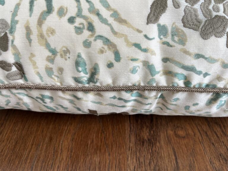 Close-up of a decorative pillow with a floral and abstract pattern in green, beige, and gray tones. The pillow has a corded seam and rests on a wooden surface.