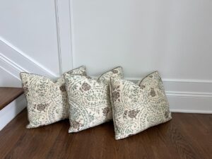 Three patterned throw pillows with a floral design are arranged on a hardwood floor near a staircase and light-colored wall.