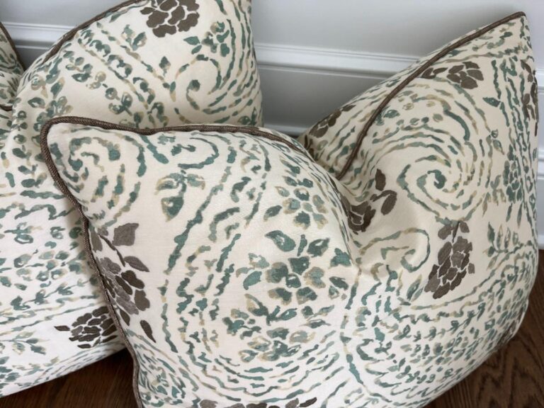 Two patterned throw pillows with an abstract floral design in shades of blue, gray, and beige. They are placed against a light-colored wall and a dark wooden floor.