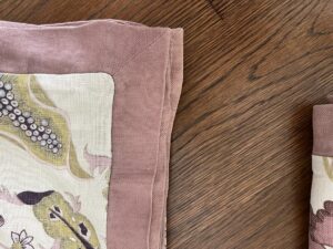 A close-up of a floral-patterned fabric piece with a pink border and a matching folded fabric piece on a wooden surface.