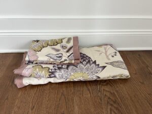 Folded floral-patterned bedspread and matching pillowcase lying on a wooden floor near a white wall.