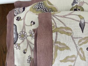 A close-up view of three folded fabric pieces with a botanical print featuring flowers and leaves, bordered by a pinkish-brown trim. The fabric is laid on a wooden surface.