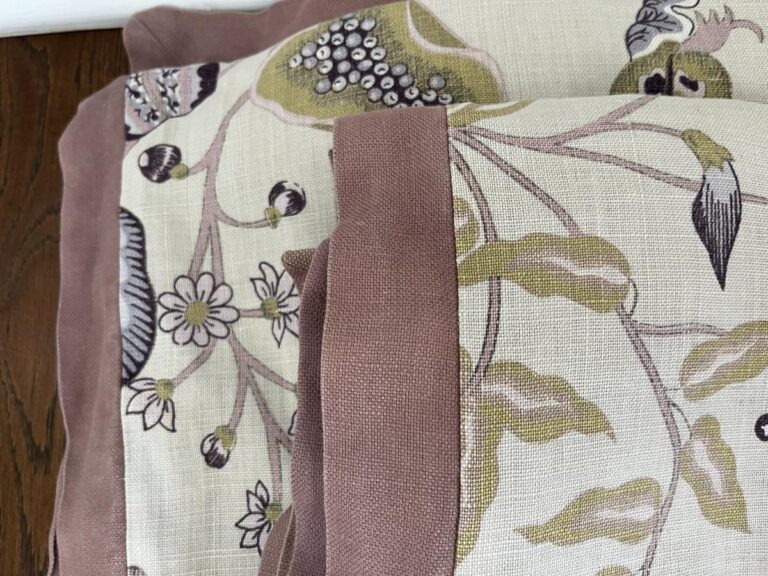 A close-up view of three folded fabric pieces with a botanical print featuring flowers and leaves, bordered by a pinkish-brown trim. The fabric is laid on a wooden surface.