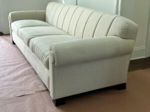 A beige, upholstered sofa with a tufted backrest and rolled arms, placed on a floor partly covered with protective paper.