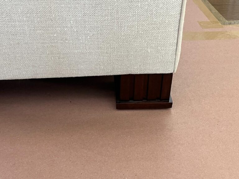 Close-up of a beige upholstered couch with a dark wooden leg on a light brown floor.