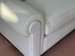 Close-up view of the arm and cushion of a light gray upholstered sofa with a welt trimming detail, positioned on a brown carpet.